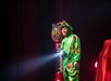 Person wearing a green dragon costume stands on stage holding a flashlight, with a gumball machine and red curtain in the background.