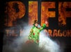 Person in a dragon costume stands on stage with "PIFF THE MAGIC DRAGON" written in the background, surrounded by smoke.