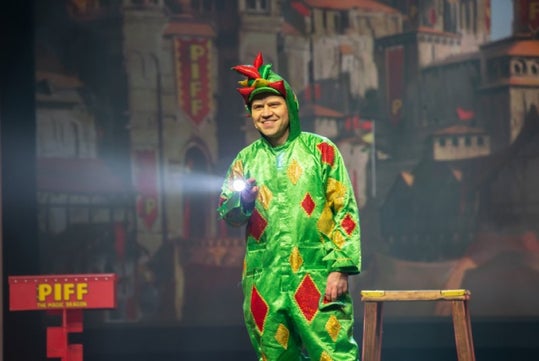 Person dressed in a green dragon costume holding a flashlight on stage, with a medieval-style backdrop and a small wooden table nearby.