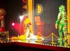 A performer in a yellow and white feathered costume sits on stage, while another in a green dragon costume stands nearby. A large gumball machine and a red background are in the scene.
