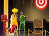 Two performers in colorful costumes, one in a yellow feathered outfit and the other in a dragon suit, prepare for a stage act involving a cannon and a target backdrop.