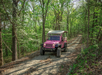 Pink jeep driving on a gravel path through a green, wooded area during the day.