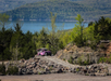 A pink off-road vehicle drives on a rocky path with a lake and green hills in the background.