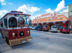 A red trolley drives down a street lined with shops and parked cars under a cloudy blue sky.