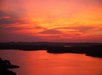 A vibrant sunset over a calm lake with silhouetted hills in the background and a small boat on the water.