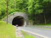 A car enters a stone-lined tunnel surrounded by green trees and foliage along a two-lane road.