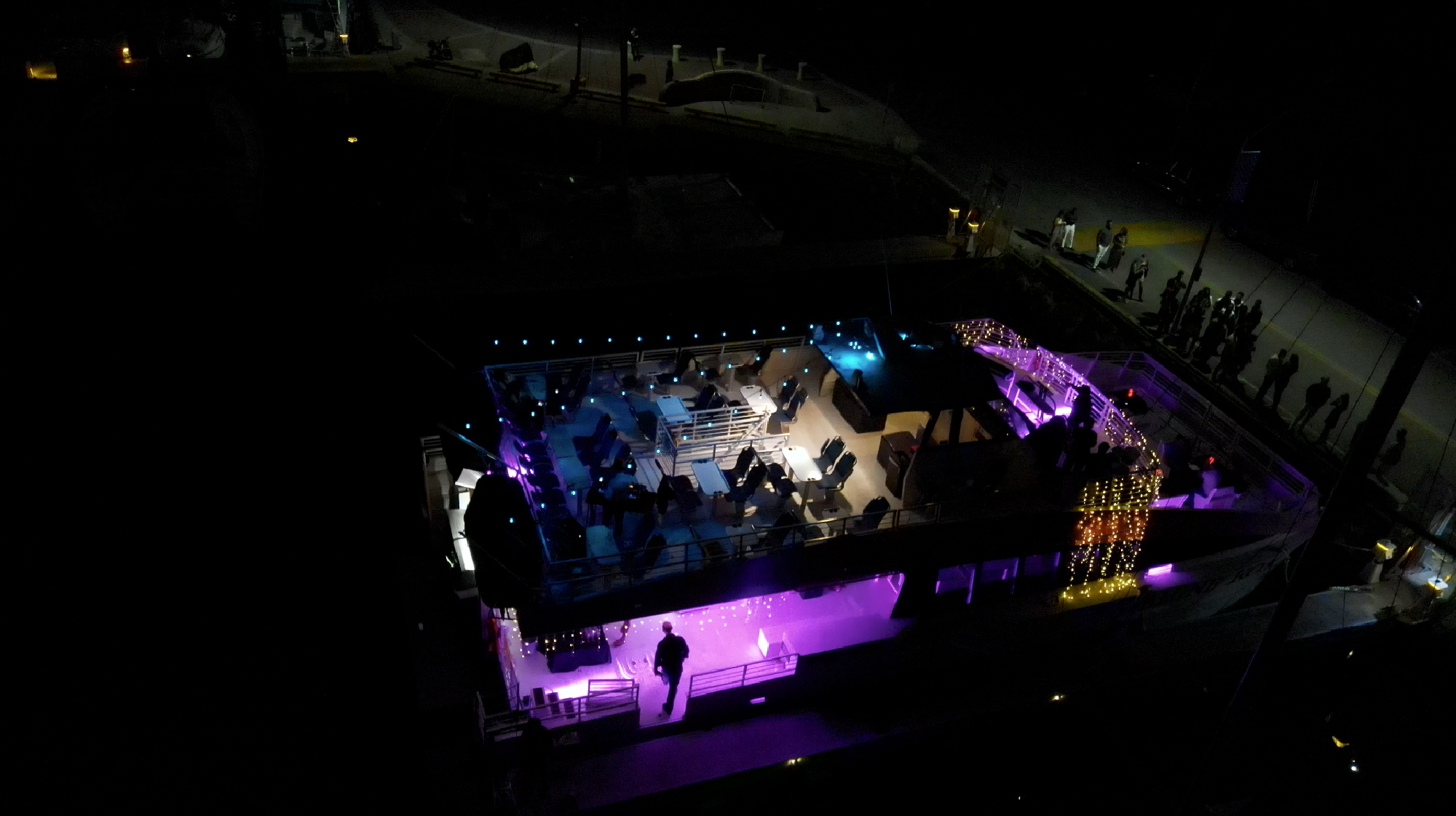 Aerial view of a docked, two-level boat at night, illuminated by purple and white lights, with empty seating areas and a few people visible on board and near the dock.
