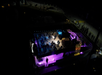 Aerial view of a yacht illuminated with purple and white lights at night. The deck has lounge seating and a figure walking.