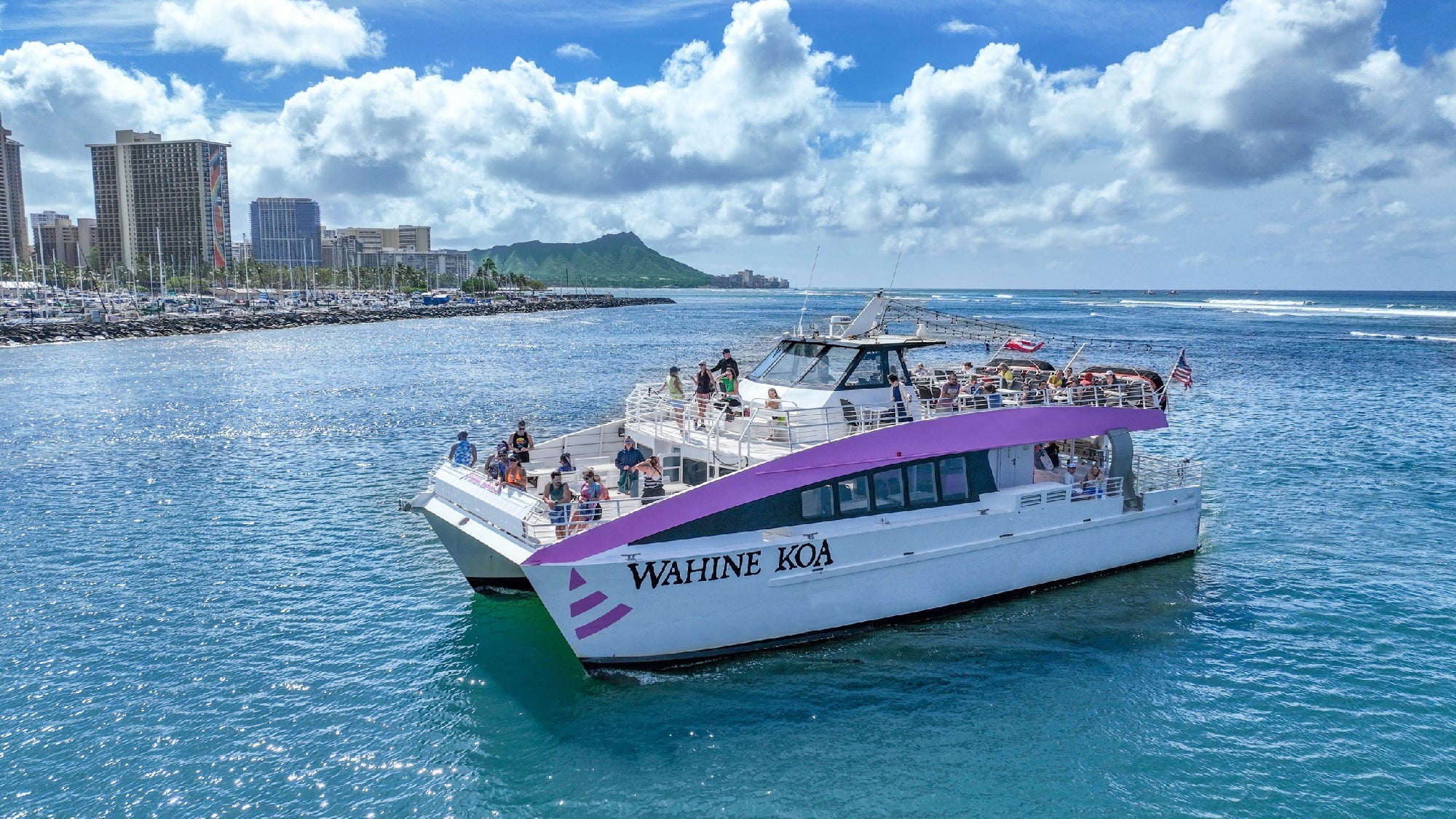 A white and purple catamaran named Wahine Koa is cruising on the ocean near a coastline with skyscrapers and a mountain in the background under a partly cloudy sky.