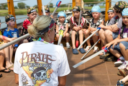 A woman in a "Pirate Adventures" shirt talks to a group of children wearing pirate hats and holding toy swords on a boat.