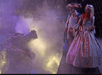 Two people in theatrical costumes face a dragon-like creature amidst fog and dramatic lighting on stage.