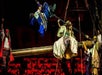 Performers in colorful costumes are suspended and staged theatrically on a large pole in front of an audience.