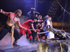 Actors in pirate costumes on stage wield swords against a crocodile prop, with a ship rigging backdrop.