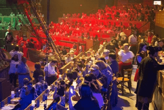 Audience seated in a dimly lit theater with a nautical theme, observing a performance on stage featuring a partial pirate ship set.