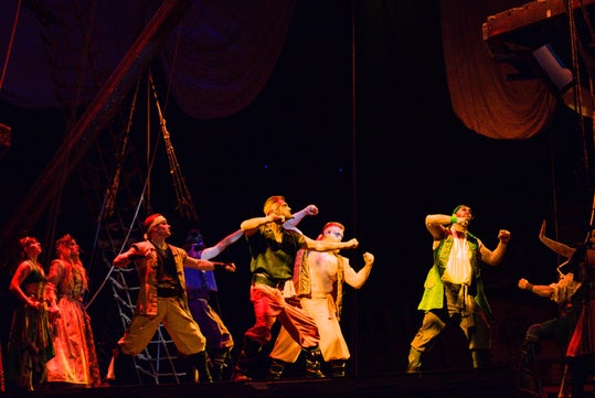 Performers dressed as pirates strike dynamic poses on a dimly lit stage resembling a ship.
