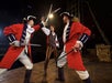 Three men in historical costumes, two in red coats with swords crossed, stand on a wooden stage with a third man in striped pants watching closely.