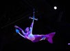 A performer dressed as a mermaid in a purple costume is suspended mid-air on an aerial apparatus during a stage show.