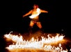 A performer in traditional attire spins a lit fire staff, with flames forming patterns on the ground in front of him against a dark background.