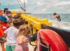 A group of children on a boat watch a person in a black vest and cap performing on the water; a red life preserver is attached to the railing.
