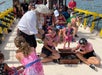 A man dressed as a pirate hands out items to children and adults on a boat; two people in striped shirts sit near an open chest filled with trinkets.