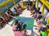 A group of children in pirate hats sit in a circle on a boat deck around a colorful treasure map, guided by two adults in striped shirts.