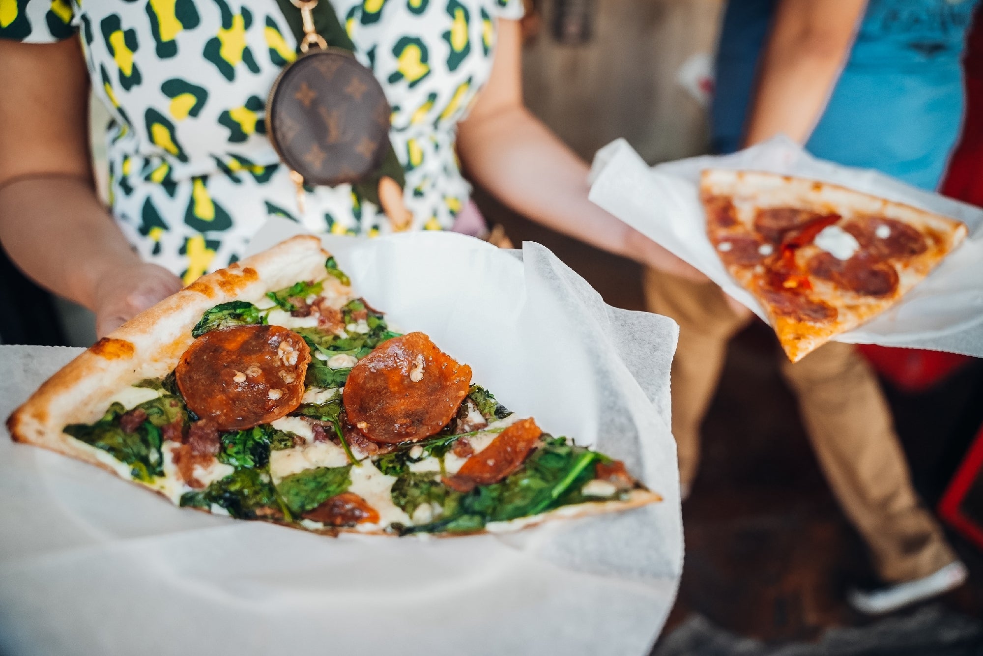 Two people hold paper plates with pizza slices topped with pepperoni, spinach, and cheese; one person wears a patterned shirt and a crossbody bag.