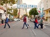 A group of people walk across a street in Little Italy, San Diego, with buildings, cars, and Italian flags in the background.