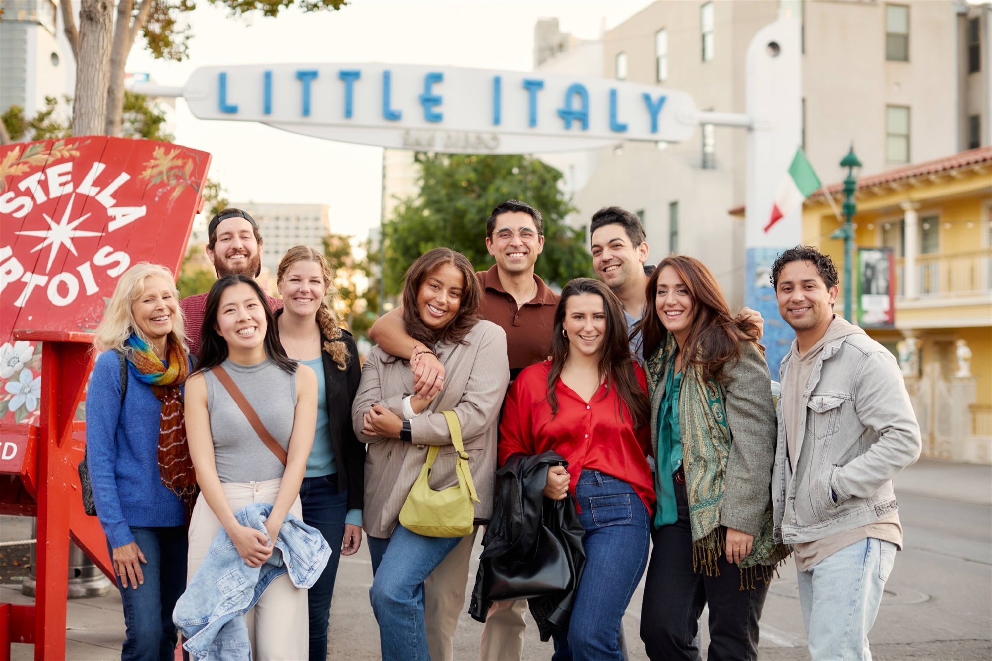 A group of ten people pose and smile for a photo under the "Little Italy" sign on a city street during the day.