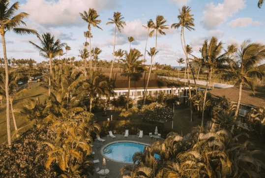 Aerial view of a tropical resort with a central swimming pool, surrounded by palm trees and lush greenery, under a partly cloudy sky.