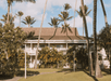 A tropical building with a brown roof is surrounded by palm trees and lush greenery under a partly cloudy sky.