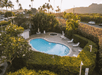 Aerial view of a small swimming pool and hot tub surrounded by hedges and palm trees, with lounge chairs around the area and mountains in the background.