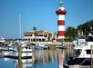 A marina with several boats docked in front of a red and white striped lighthouse and a building under a clear blue sky.