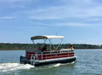 A red pontoon boat with a canopy travels on a calm body of water under a clear blue sky. Three people are on board, and distant greenery is visible in the background.