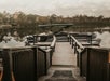 A wooden dock leads to a pontoon boat moored on a calm lake, with trees and cloudy skies reflected in the water.
