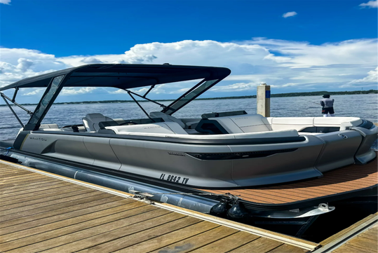 A gray and white pontoon boat is docked at a wooden pier on a sunny day, with water and clouds visible in the background.