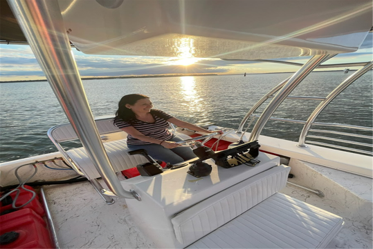 A woman wearing a striped shirt steers a boat on calm water during sunset, with sunlight reflecting on the water's surface.