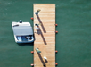 Aerial view of a dock extending into green water with a small boat moored to the left side. Shadows from posts fall across the wooden dock.