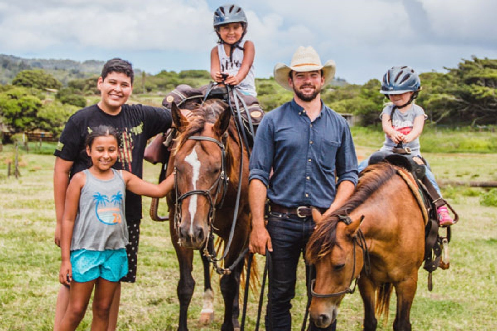 A group of people, including three children and an adult, stand with two horses in a grassy field. The children and adult are wearing helmets, and the adult has a cowboy hat.