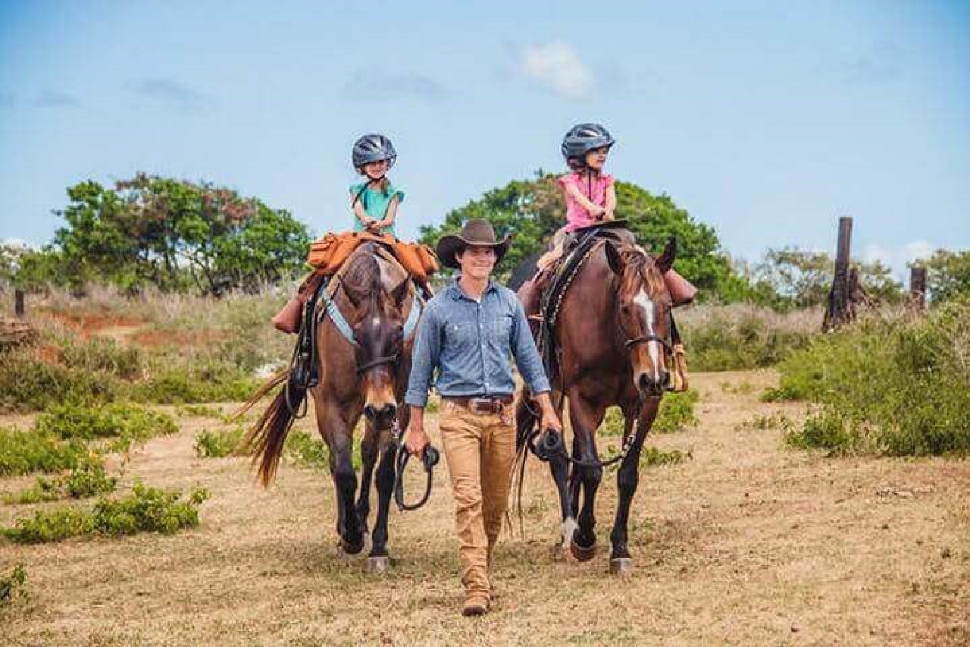 A person leads two horses, each ridden by a child wearing a helmet, on a grassy path under a blue sky.