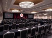 A large conference room with rows of black tables lined with black chairs in front of a podium and two large projector screens.