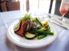 A plate of sliced beef served with green beans, zucchini, yellow squash, and carrots on a white tablecloth, with a glass of water and a pink drink in the background.