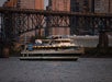 A white and green passenger boat travels on a river with a city skyline and a metal bridge in the background at dusk.
