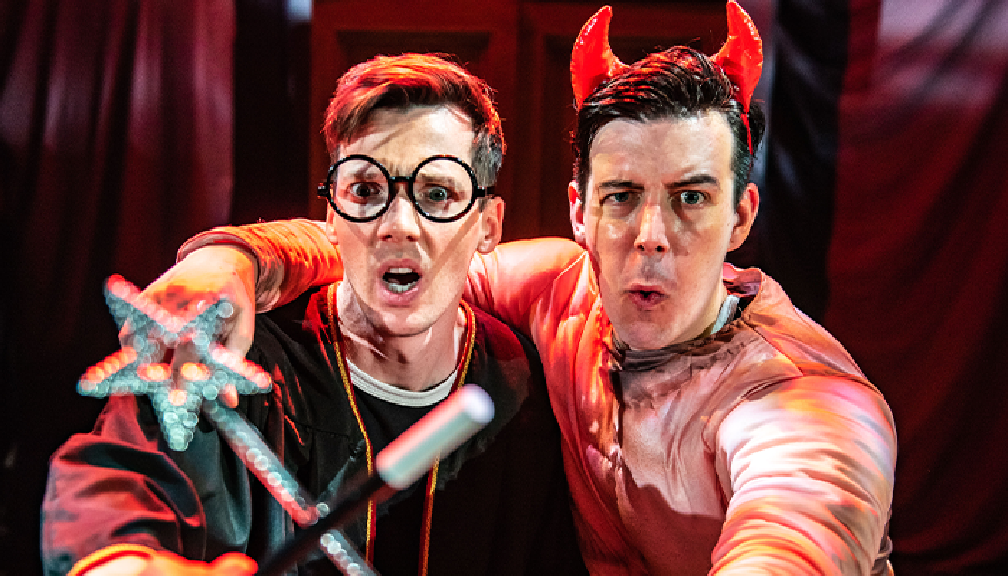 Two men in costume pose for the camera; one wears round glasses and a robe, the other wears a red devil costume with horns.