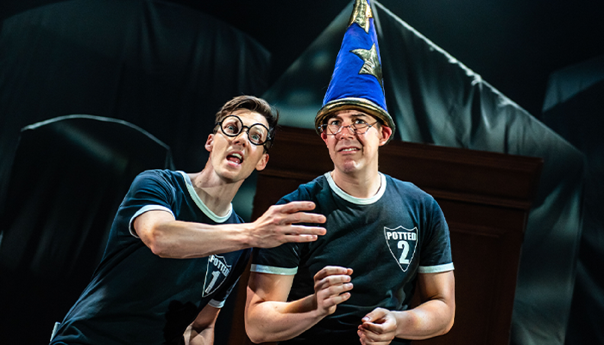 Two people in matching shirts, one wearing a wizard hat and the other wearing round glasses, perform on stage with dramatic expressions.