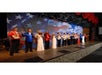A group of performers in colorful costumes stand on stage in front of a large American flag backdrop, posing and smiling at the audience.
