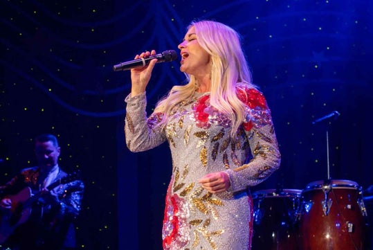 A woman in a sparkly, floral dress sings into a microphone on stage; a musician with a guitar and conga drums are visible in the background under stage lighting.
