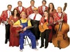 Ten people in coordinated red outfits pose with musical instruments, including guitars, a banjo, and a double bass; two people in costume sit at the front.