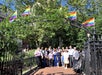 A group of people stand under an iron archway decorated with Pride flags in a sunny, leafy outdoor setting.