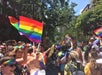 A large crowd gathers outdoors in daylight, waving rainbow pride flags and celebrating during a parade on a city street lined with trees and buildings.