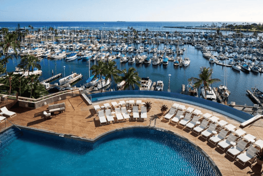 Aerial view of a marina with numerous boats docked. In the foreground, a resort pool area with lounge chairs overlooks the water, set against a backdrop of blue sky and ocean.